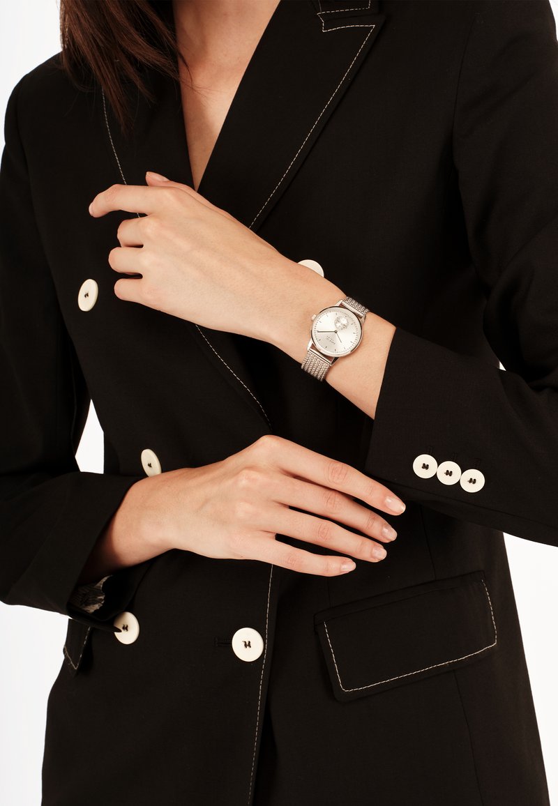 Montre en argent avec un cadran rond et un bracelet en maille, portée avec un blazer noir doté de boutons clairs et de coutures contrastées.