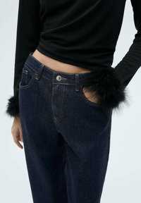 Tmavomodré džíny vyrobené z denimu s vysokým pasem a bočními kapsami, s černými kožešinovými detaily na manžetách. Hladká textura a robustní design.