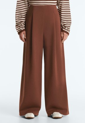 Szerokie brązowe spodnie noszone z długim rękawem, brązowo-białą bluzką w paski oraz białymi butami, pokazane od pasa do stóp.