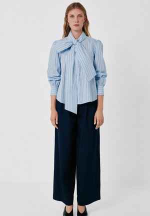 Femme debout portant un chemisier rayé bleu avec un grand nœud et un pantalon large bleu marine sur un fond blanc uni.