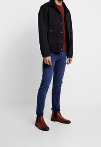 Svart denimjacka med shearlingkrage, röd skjorta under, blå slim-fit jeans och bruna läderankelboots. Modell som står på sidan.
