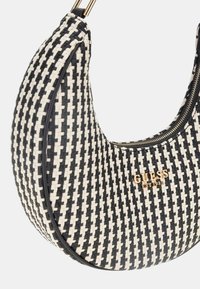 Sac à épaule tissé noir et blanc avec une forme incurvée, accents en matériel doré et une fermeture éclair affichant le nom de la marque "GUESS" de manière proéminente.