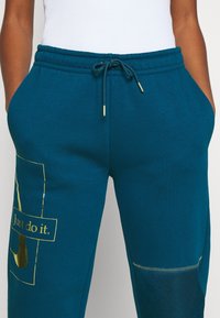 Pantalones de chándal en color verde azulado con cintura elástica, detalles en dorado y logo impreso. Incluye bolsillos laterales y un parche en la pierna izquierda.