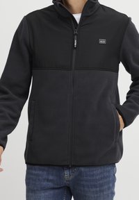 Chaqueta de forro polar negra con una sección superior lisa, cierre de cremallera y bolsillos laterales. La tela parece suave y texturizada. Presenta un emblema en el pecho.