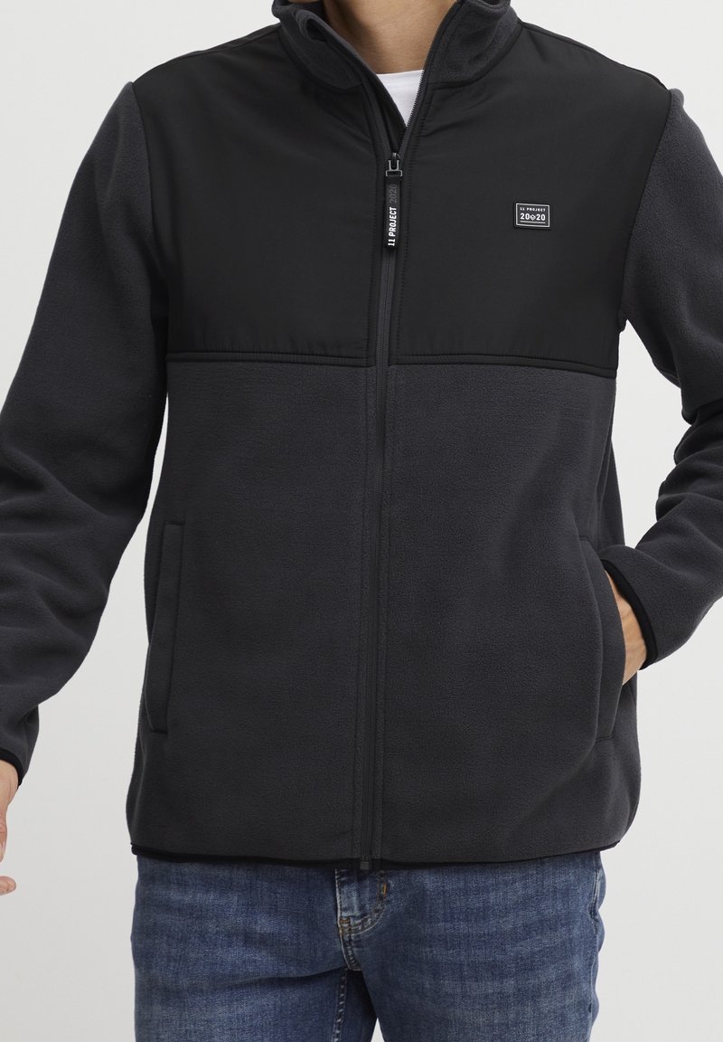 Chaqueta de forro polar negra con una sección superior lisa, cierre de cremallera y bolsillos laterales. La tela parece suave y texturizada. Presenta un emblema en el pecho.