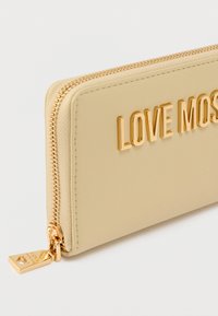 Πορτοφόλι μπεζ με ομαλή υφή, με χρυστό φερμουάρ και ανάγλυφη επιγραφή "LOVE MOST" στο μπροστινό μέρος.