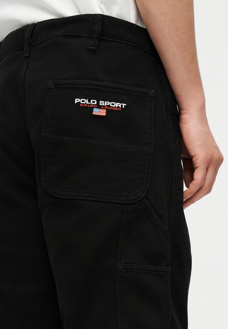Poche d'un pantalon noir avec le logo blanc et rouge "Polo Sport Ralph Lauren" et une petite broderie du drapeau américain à l'arrière, porté par une personne.
