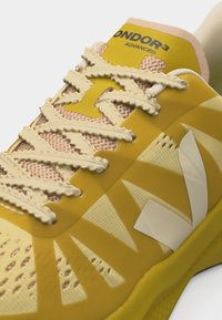 Κοντινό πλάνο ενός κίτρινου αθλητικού παπουτσιού με υφασμένες κορδόνια, αναπνεύσιμο δίχτυ και λευκό λοξό λογότυπο στο πλάι.