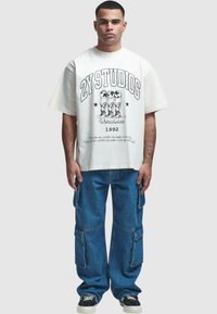 Balta per didelis T-shirt su juodu grafiniu atspaudu ir tekstu. Mėlynos džinsinės kargo kelnės su šoninėmis kišenėmis. Juodi sportbačiai su baltais padais.