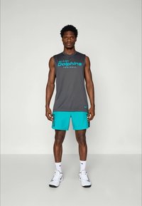 T-shirt sans manches grise avec le texte "Miami Dolphins Football", associée à un short turquoise et des baskets blanches. Design athlétique simple.