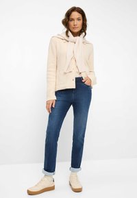 Cardigan a maglia color crema con una sciarpa annodata, abbinato a jeans blu scuro e sneakers beige. Presenta polsini arrotolati e tasche laterali.