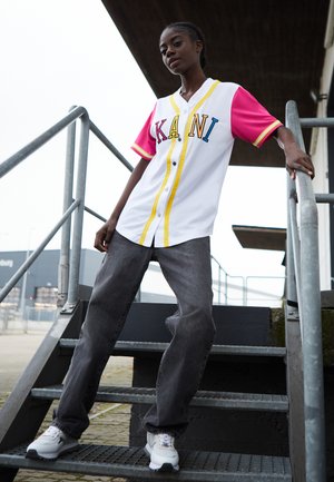 Weißes Trikot mit rosa Ärmeln, gelben Akzenten und "KANI" in blauen Buchstaben. Getragen mit lockeren grauen Jeans und weißen sportlichen Sneakern.