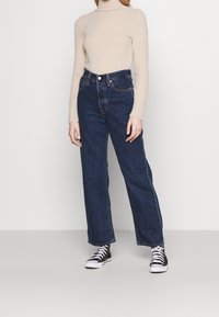 Jersey de cuello vuelto de color beige claro combinado con jeans anchos de talle alto en azul oscuro y zapatillas negras y blancas. Textura suave, ajuste casual.