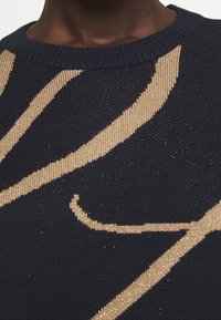 Stickad tröja i marinblått med ett strukturerat, guldtråds växtmönster. Rund halsringning; mjukt, elastiskt tyg med en subtil lyster.