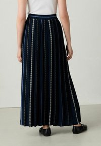 Gonna plissettata blu navy con righe verticali argento e oro, dotata di una vita e un design lungo. Indossata con ballerine nere.