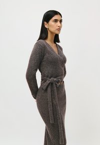 Kvinde med lige sort hår iført en brun strikket omslagstrøje og matchende nederdel med bånd, stående foran en ensfarvet lys baggrund.