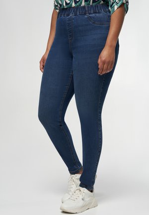 Leggings en denim bleu foncé avec une taille élastique, présentant une texture lisse et une coupe skinny, associées à des chaussures de sport blanches.