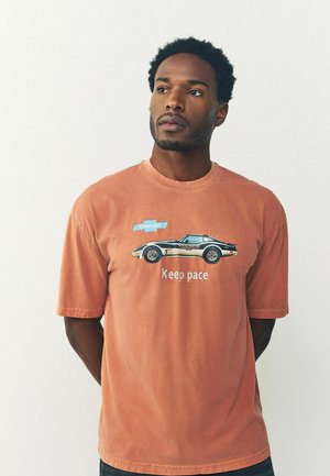 Short-sleeve, rust-colored t-shirt made of cotton. Features a graphic of a car and text "Keep pace." Casual fit and smooth texture.
