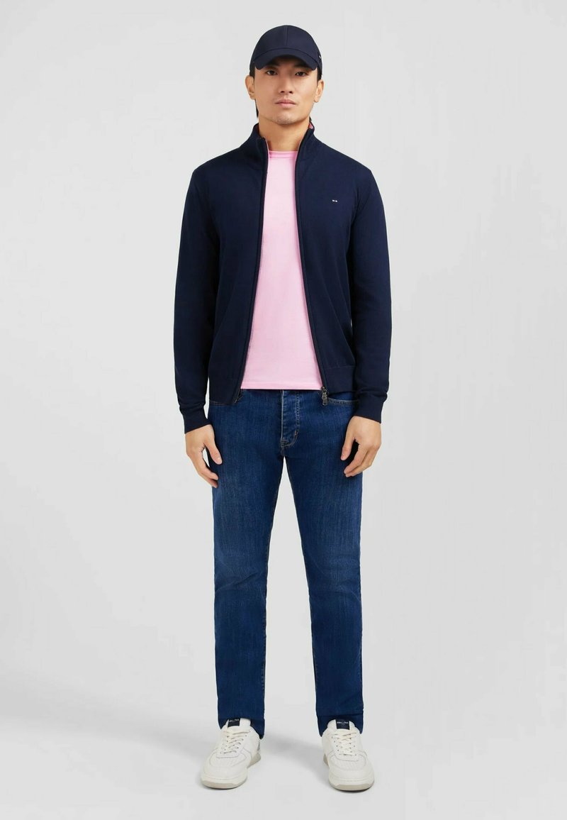 Veste zippée navy sur un t-shirt rose, associée à un jean bleu et des baskets blanches. Le modèle porte une casquette navy. Design simple sans motifs.