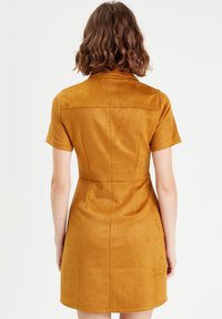 Robe à manches courtes en suédine moutarde, corsage ajusté avec une jupe évasée, coutures arrière et détails de pattes de boutonnage. Texture lisse sans motifs visibles.