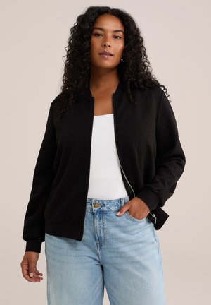 Femme aux cheveux bouclés portant une veste noire zippée, un haut blanc et un jean bleu clair, debout avec une main dans la poche.