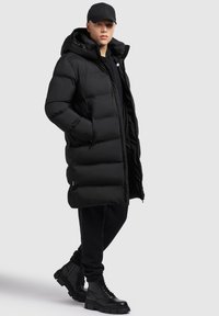 Sort pufferjakke med høj krave og hætte, med quiltet design og to sidelommer, sammen med sorte bukser og solide sorte støvler.