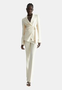 Completo sartorial crema con giacca avvolgente dotata di cintura, abbinato a pantaloni tapered. Tessuto liscio con sottili linee verticali, completato da sandali con apertura sulle dita.