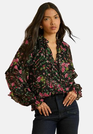 Black floral blouse featuring a ruffled neckline and cuffs, adorned with pink and green flowers on a sheer fabric. Worn with dark jeans.