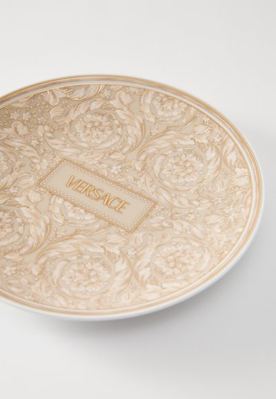 Okrągły porcelanowy talerz z misternym złotym kwiatowym wzorem i centralną etykietą "VERSACE" w ozdobnej ramce. Delikatne beżowe i białe kolory.