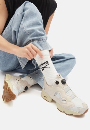 Bēša Reebok sporta apavi ar teksturētu augšdaļu, izgriezumu dizainu un biezām zolēm. Valkāti ar baltām zeķēm ar melnu logotipu, kopā ar džinsa biksēm.