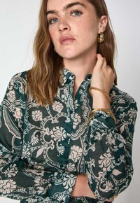 Camicia a maniche lunghe con motivo floreale verde, dotata di bottoni e colletto. Braccialetti d'oro indossati al polso.