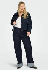 Temno denim jakna z dvema žepoma na prsih in srebrno opremo, kombinirana z visokim pasom temnih kavbojk z zavihki na robu in rdečimi šivnimi poudarki.