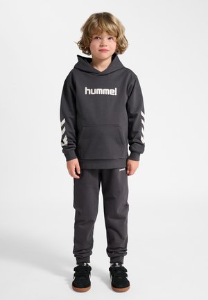 Ung dreng iført en sort hummel-hoodie med chevronstriber på ærmerne, matchende sorte joggingbukser og sorte sneakers, stående på hvid baggrund.