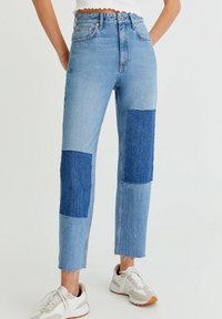 Jean denim bleu taille haute avec des accents de patch bleu plus foncé sur les genoux, ourlet effiloché, et coupe droite. Associé à des baskets blanches.