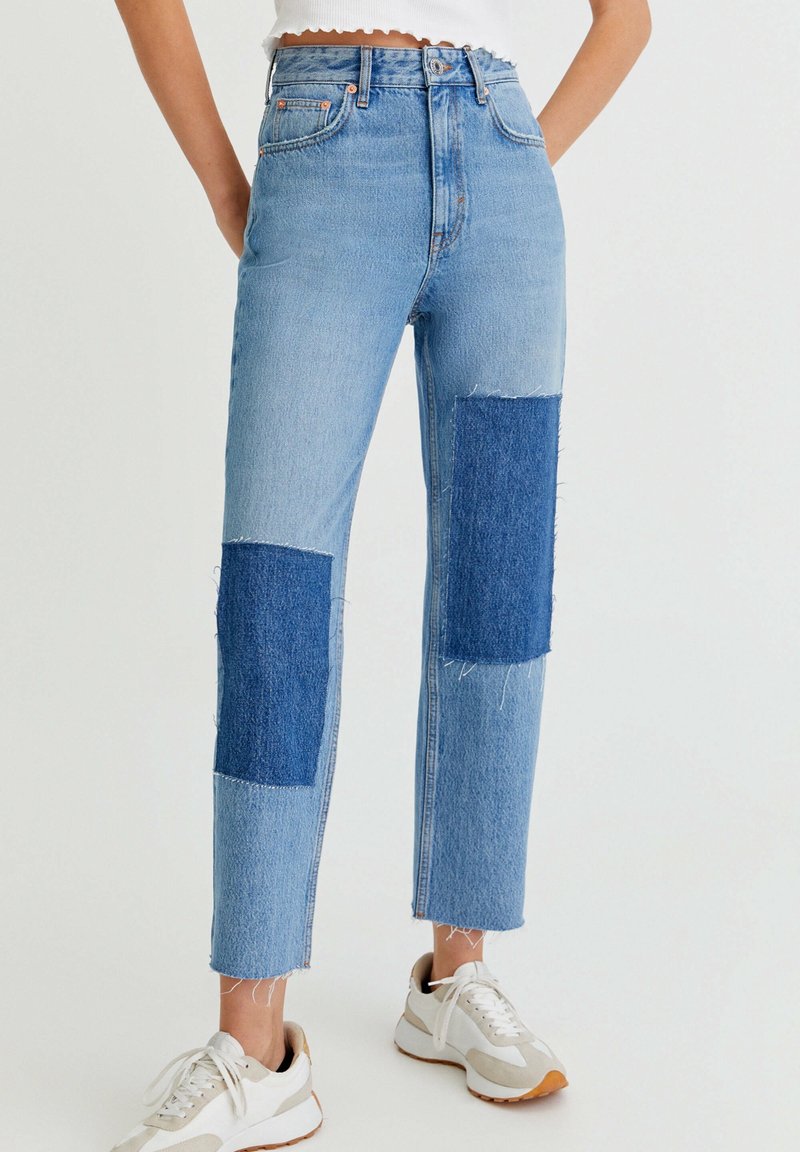 Jean denim bleu taille haute avec des accents de patch bleu plus foncé sur les genoux, ourlet effiloché, et coupe droite. Associé à des baskets blanches.