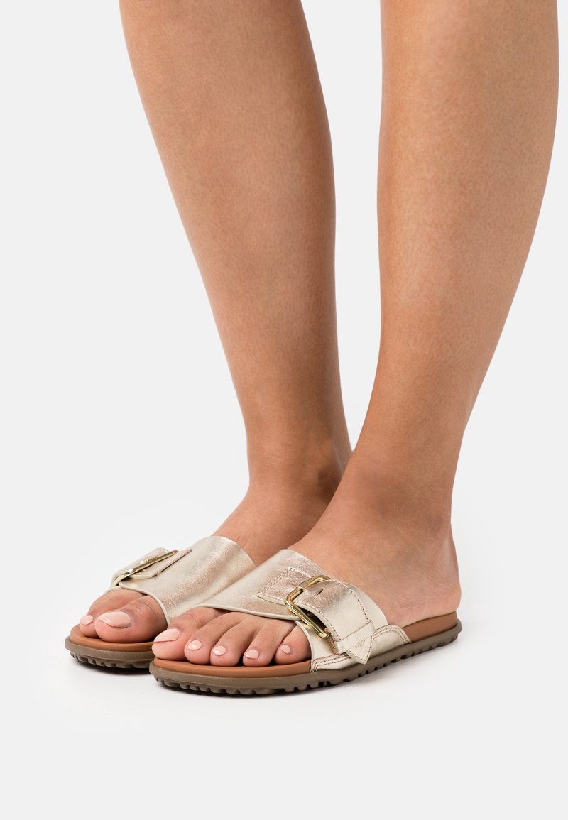 Pieds portant des sandales à glissière métalliques beige avec larges brides à boucle et semelles brunes texturées sur un fond clair.