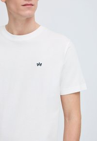 Hvit bomulls t-skjorte med korte ermer, rund hals, med en liten brodert logo i marineblått på brystet. Glatt tekstur.