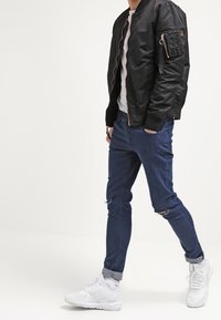 Osoba ubrana w czarną kurtkę bomberską, białą koszulę, podarte niebieskie skinny jeansy i białe trampki, stojąca z jedną ręką w kieszeni.