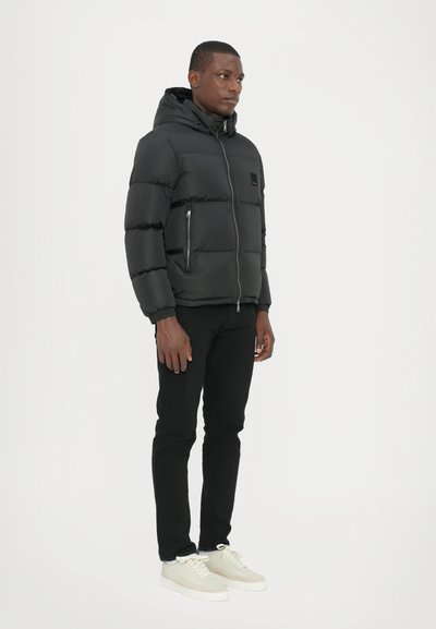Doudoune noire avec capuche, présentant des coutures horizontales, une fermeture éclair à l'avant et des poches latérales. Associée à un pantalon noir et des baskets légères.