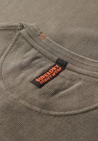 Grijze gestructureerde stof met een ronde halslijn, voorzien van een zwart label met oranje tekst, "SUPERDRY UTILITY WEAR," nabij de kraag.