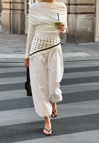Top bianco a maniche lunghe con arricciature, abbinato a pantaloni bianchi larghi. Accentuato con una cintura a pois. Tiene in mano una bevanda verde e una borsa piccola.