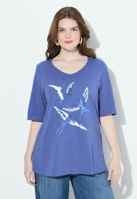 Mujer con cabello castaño ondulado que lleva una camisa de manga corta azul con un gráfico de pájaro y jeans azules, de pie frente a un fondo blanco.