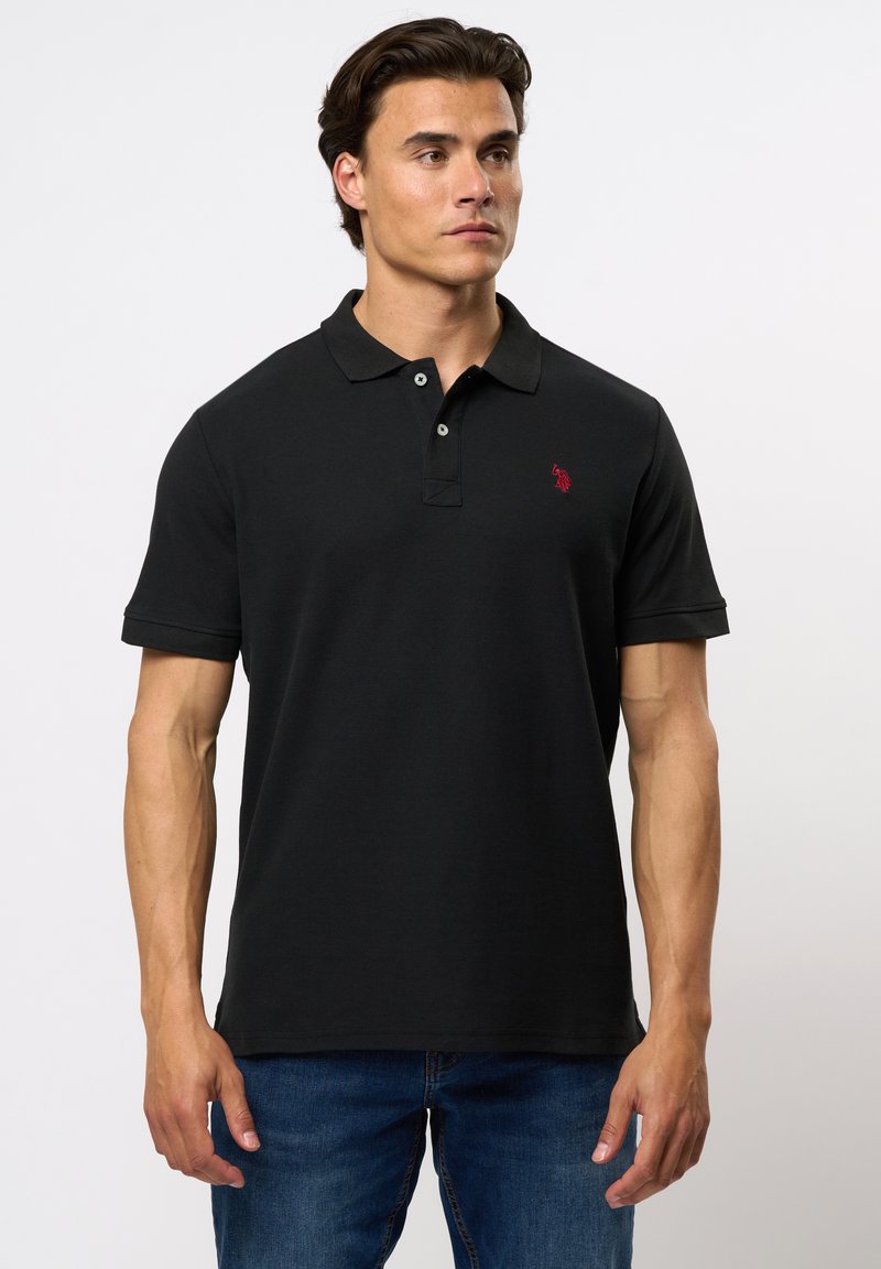 Zwarte poloshirt met ribbelstructuur, drie knoopjes aan de voorkant en een rode logo-accent op de linkerborst. G gedragen met blauwe spijkerbroek.