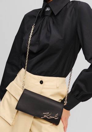 Borsa a pochette in pelle nera con tracolla a catena, caratterizzata da dettagli del nome in rilievo in argento. Texture liscia, forma rettangolare, design minimalista.