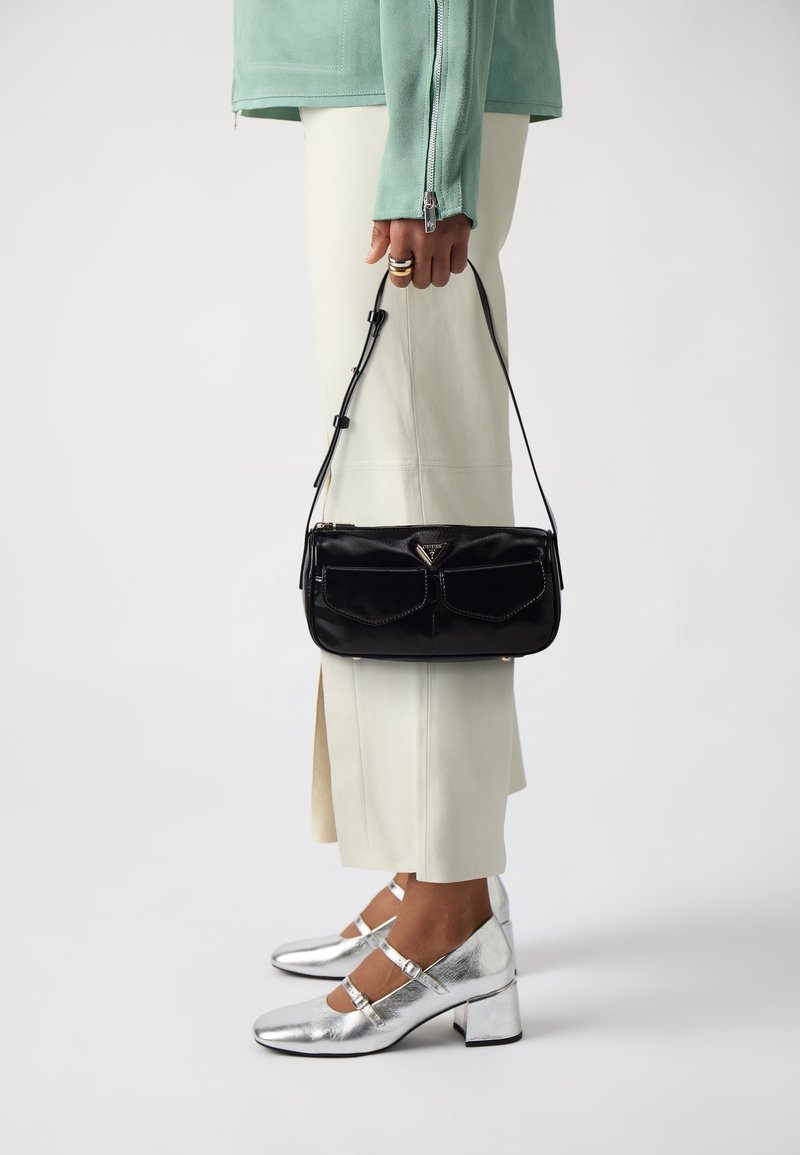 Personne tenant un sac à main noir, portant une jupe blanche, une veste verte menthe et des chaussures Mary Jane argentées à petit talon, debout sur fond blanc.