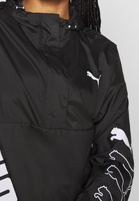 Svart vindjacka i lätt material, med en delvis dragkedja och vita Puma-loggor på ärmarna och framsidan.
