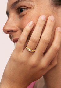 Bague en or avec une surface texturée, mettant en valeur une pierre précieuse ovale centrale, portée sur une main touchant le visage. Teinte de peau lisse, fond neutre.