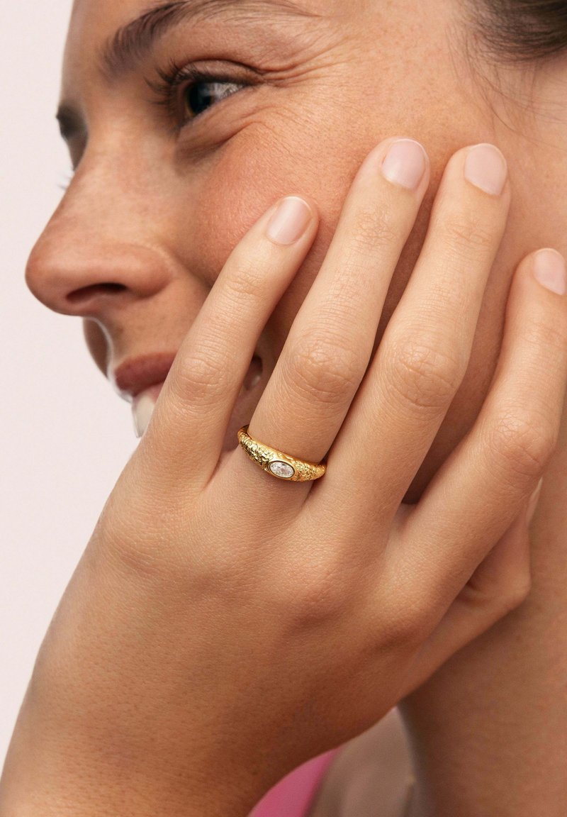Bague en or avec une surface texturée, mettant en valeur une pierre précieuse ovale centrale, portée sur une main touchant le visage. Teinte de peau lisse, fond neutre.