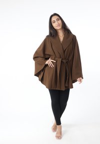 Abrigo envolvente marrón con un amplio cuello, cintura ceñida con cinturón y mangas fluidas, combinado con leggings negros y sandalias transparentes. Textura suave.