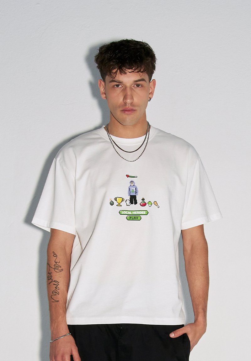 Biała bawełniana koszulka z grafiką w stylu pixel art, na której znajduje się postać, trofea i przedmioty, a także napis "LOKALNI BOHATEROWIE GRAJĄ" w kolorze zielonym.
